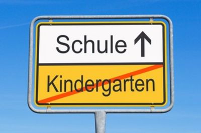 Das Bild zeigt ein Verkehrsschild (Ortsausgangsschild). In der unteren Hälfte des Schildes steht das durchgestrichene Wort Kindergarten. In der oberen Hälfte steht das Wort Schule mit einem nach oben zeigenden Pfeil.