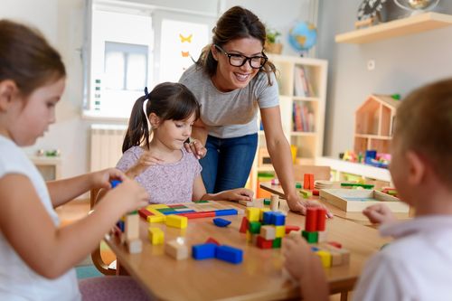Das Bild zeigt eine Erzieherin und drei Kinder in einer Kindertageseinrichtung an einem Tisch. Die Kinder bauen mit bunten Holzklötzen.