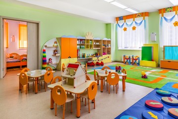 Das Bild zeigt ein Spielzimmer in einer Kindertageseinrichtung. Es sind bunte Kindermöbel (u. a. Tische, Stühle und Schränke), Spielsachen und Teppiche zu sehen.